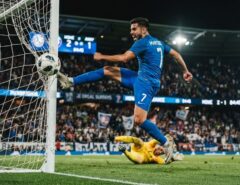 A male soccer player kicking a soccer ball into the goal net during a stadium match.